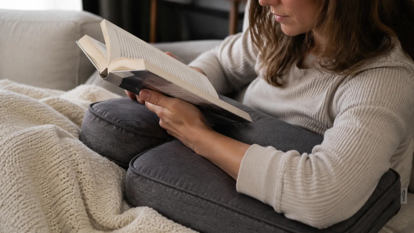 Close-up of a person's forearms resting comfortably on the padded arms of a reading pillow while holding a book